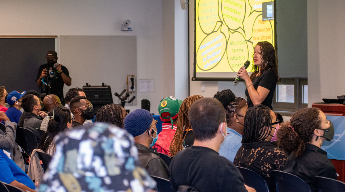 Woman standing up front of a classroom speaking into a mic with a group of people seated in front of her 